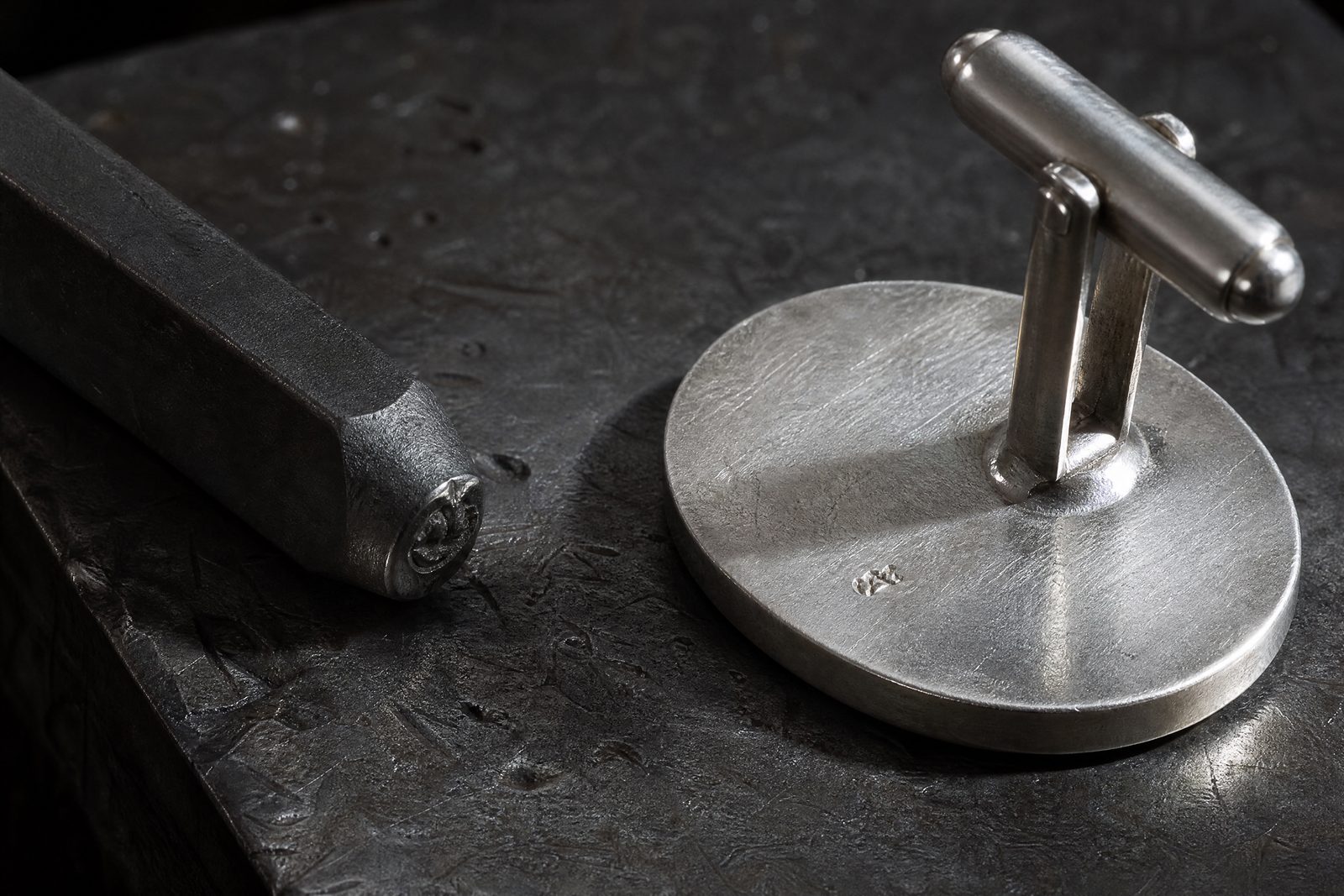 A steel hallmarking punch resting on a scarred steel bench block beside the reverse face of a silver cufflink.